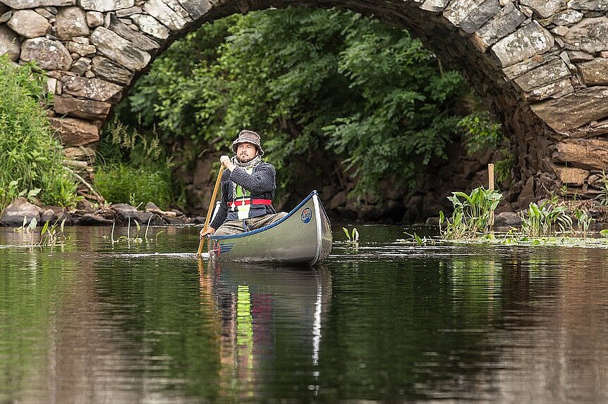 Dieses Bild zeigt einen Mann rudernd im Kanu vor einer Steinbrücke.