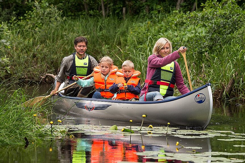 Dieses Bild zeigt eine Familie mit Vater, einem kleinen Mädchen, einem kleinen Jungen und der Mutter, die im Kanu eine Flusslandschaft entlang paddeln.