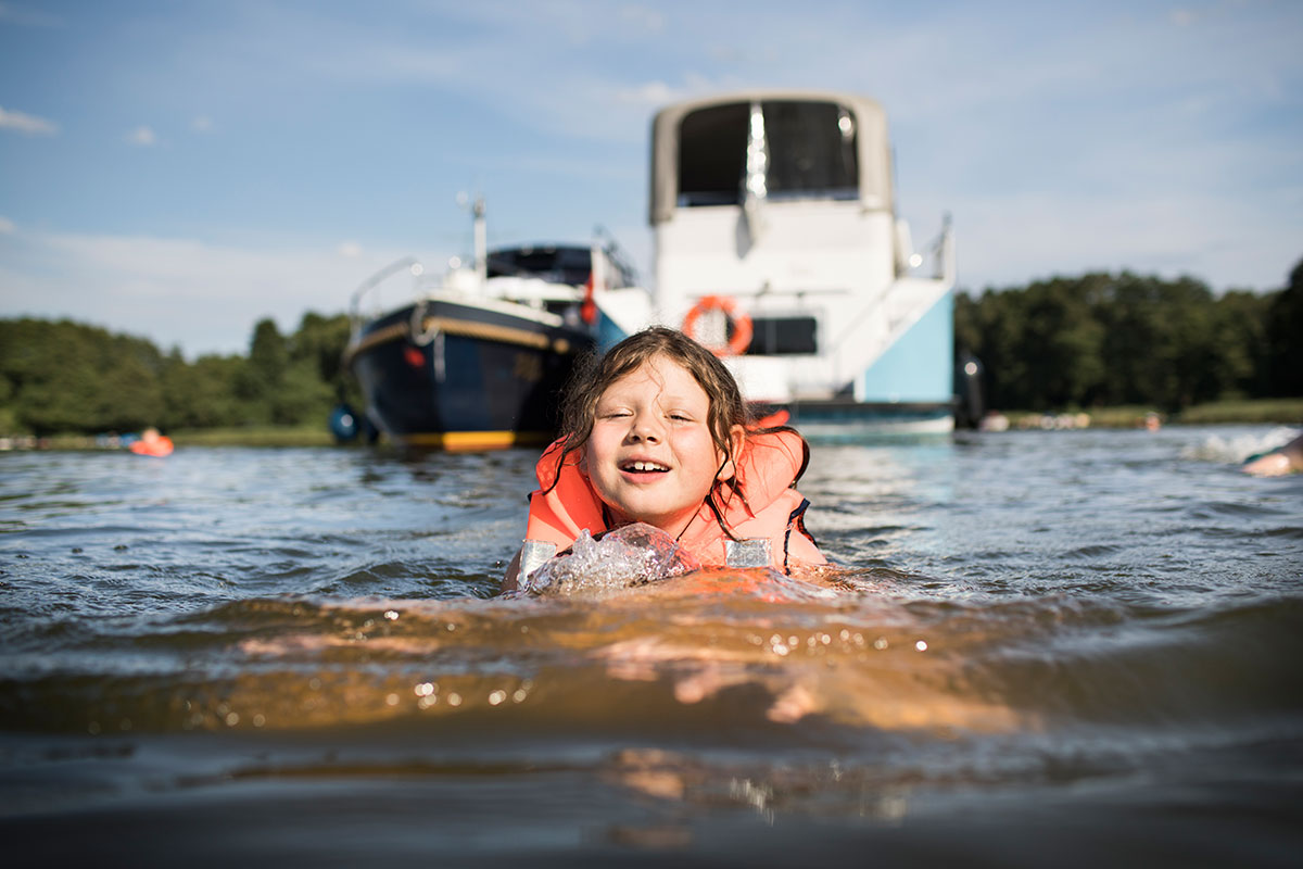 Ein Kind schwimmt in der Müritz, im Hintergrund liegen 2 Yachten vor Anker.